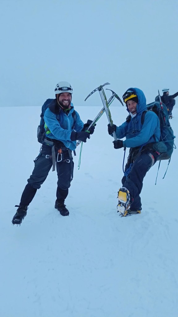 Dos montañistas en una cumbre con el equipo de alpinismo, crampones, piolet, casco, gafas, linterna, arnés, mochilas y otros.